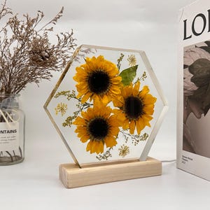 Pu&ograve; includere: Un blocco esagonale in resina trasparente contenente tre girasoli gialli pressati e piccolo fogliame verde, montato su una base rettangolare in legno chiaro. L'oggetto presenta una finitura liscia e trasparente che mette in risalto le texture.