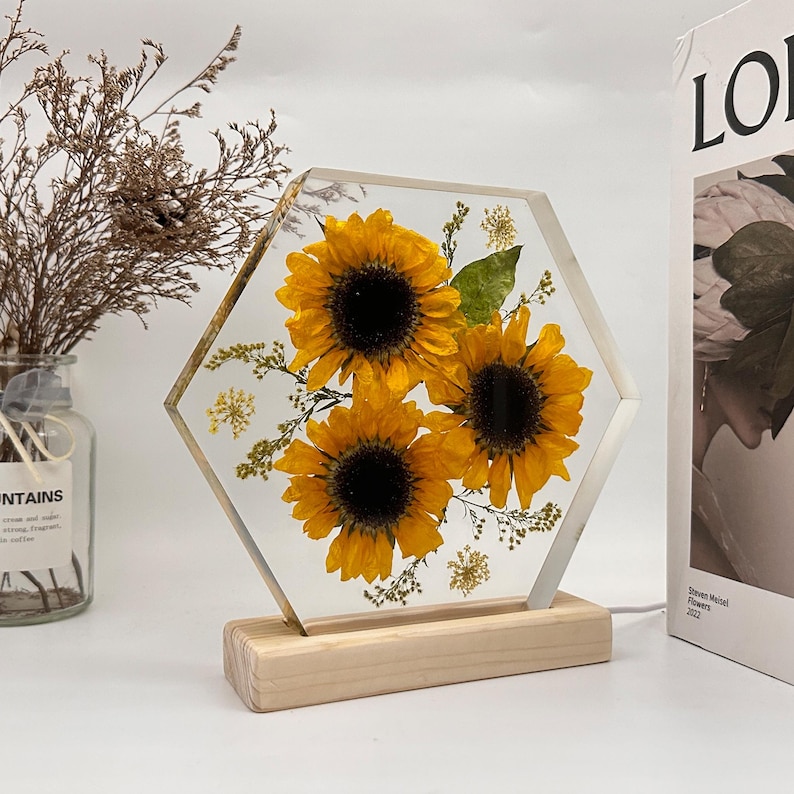 Pu&ograve; includere: Un blocco esagonale in resina trasparente contenente tre girasoli gialli pressati e piccolo fogliame verde, montato su una base rettangolare in legno chiaro. L'oggetto presenta una finitura liscia e trasparente che mette in risalto le texture.