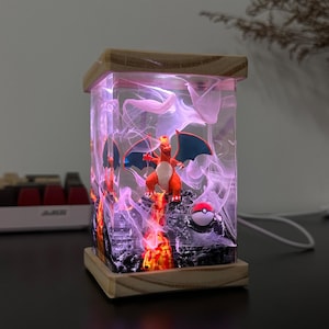 K&ouml;nnte beinhalten: Eine klare Harzlampe mit einer roten und orangefarbenen Charizard-Pok&eacute;mon-Figur im Inneren. Die Lampe leuchtet in einem rosafarbenen Licht und hat einen wei&szlig;en Raucheffekt. Am Boden der Lampe befindet sich ein roter und wei&szlig;er Pok&eacute;ball.
