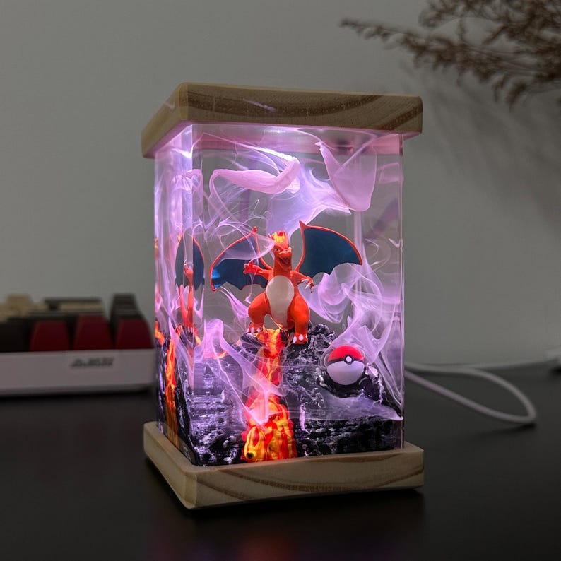 K&ouml;nnte beinhalten: Eine klare Harzlampe mit einer roten und orangefarbenen Charizard-Pok&eacute;mon-Figur im Inneren. Die Lampe leuchtet in einem rosafarbenen Licht und hat einen wei&szlig;en Raucheffekt. Am Boden der Lampe befindet sich ein roter und wei&szlig;er Pok&eacute;ball.