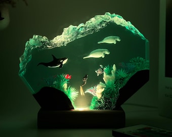 May include: A decorative lamp depicting an underwater scene with a black and white orca, manatees, and a diver. The lamp has a green glow, with a textured, wave-like top and a wooden base. The scene includes coral and other aquatic life.