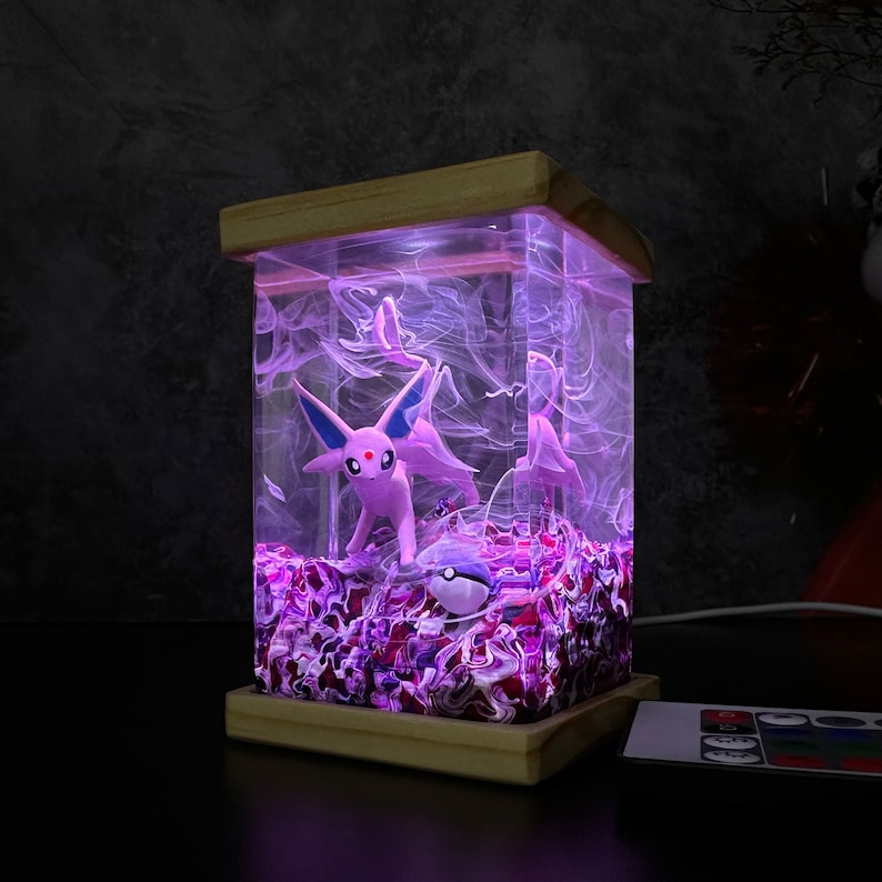 May include: A pink and blue Pokemon figurine of Espeon is encased in a clear resin cube with a swirling purple and white pattern. The cube is lit from within with a purple light and sits on a wooden base.