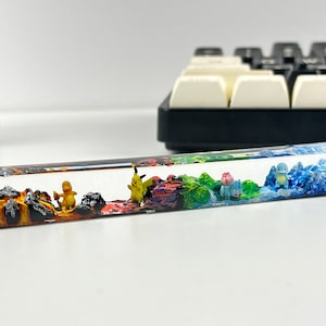 Może przedstawiać: Przezroczysty klawisz z żywicy z wielokolorowym wzorem krajobrazu przedstawiającym miniaturowe figurki postaci Pokemon&oacute;w. Krajobraz obejmuje obszar wulkaniczny, obszar trawiasty i obszar wodny.