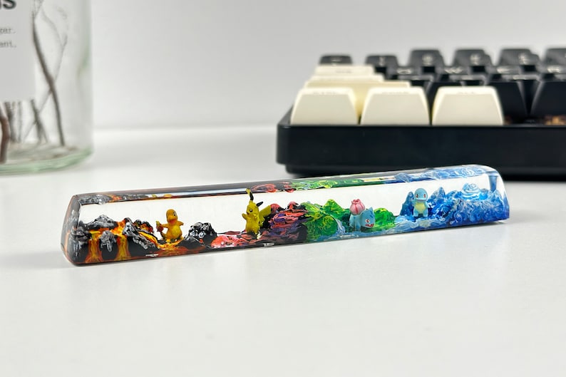Może przedstawiać: Przezroczysty klawisz z żywicy z wielokolorowym wzorem krajobrazu przedstawiającym miniaturowe figurki postaci Pokemon&oacute;w. Krajobraz obejmuje obszar wulkaniczny, obszar trawiasty i obszar wodny.