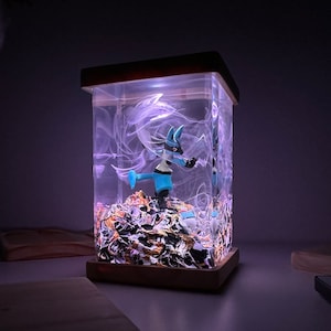 May include: A blue and white Pokemon figurine is encased in a clear resin cube with a wooden base. The cube is illuminated from within, creating a glowing effect. The resin contains swirling white and gold accents.