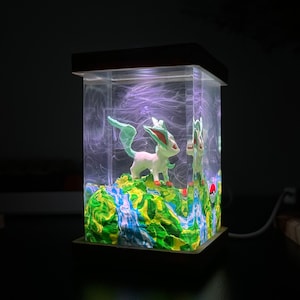 May include: A clear glass cube with a wooden base, containing a green and white figurine of a Pokemon character, standing on a green and blue painted landscape. The cube is lit from the inside, creating a glowing effect.