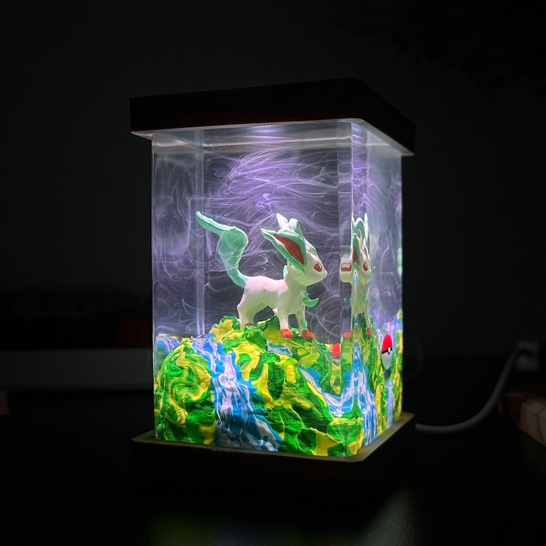 May include: A clear glass cube with a wooden base, containing a green and white figurine of a Pokemon character, standing on a green and blue painted landscape. The cube is lit from the inside, creating a glowing effect.