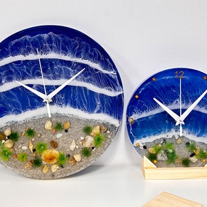 Könnte beinhalten: Zwei Uhren aus Harz mit Strandmotiv. Die größere Uhr ist blau mit weißen Wellen und einem Sandstrand mit Muscheln. Die kleinere Uhr steht auf einem Holzsockel.
