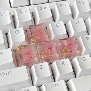 K&ouml;nnte beinhalten: Eine wei&szlig;e Tastatur mit rosa und goldenen Blumen-Tastenkappen. Die Tastenkappen bestehen aus klarem Harz mit echten Blumen und goldenen Flocken, die darin eingebettet sind.