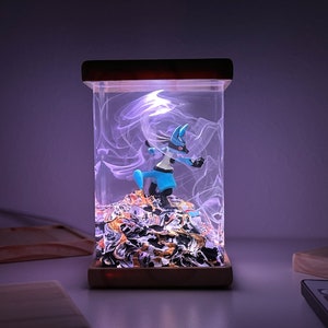 May include: A blue and black Pokemon figurine of Lucario is inside a clear glass lamp with a wooden base. The lamp is lit up with a white light, creating a swirling effect inside the glass.