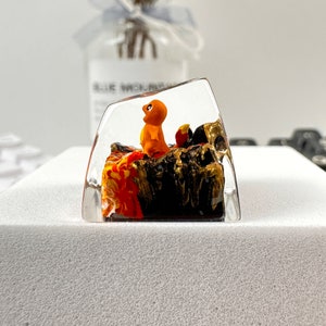May include: A clear resin keycap with a small orange and yellow figurine of a Pokemon character standing on a black and gold base with red and orange flames.