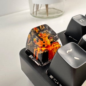 May include: A black and orange keycap with a clear resin top. The keycap features a small orange creature with a yellow tail and a black background. The keycap is sitting on a black keyboard.