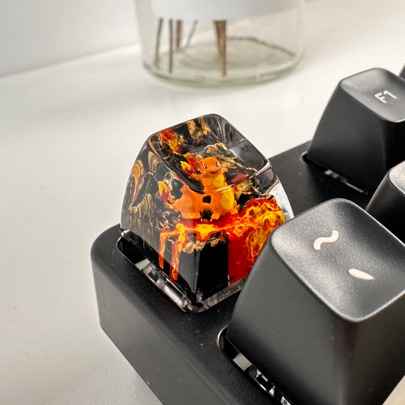May include: A black and orange keycap with a clear resin top. The keycap features a small orange creature with a yellow tail and a black background. The keycap is sitting on a black keyboard.