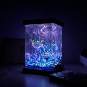 May include: A blue and white Pokemon character figurine is inside a clear resin lamp with a blue and white swirling base. The lamp is on a dark surface and is lit up with a blue light.