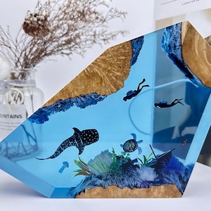 Könnte beinhalten: Eine blaue Harzskulptur, die eine Ozeanszene mit einem Walhai, einer Schildkröte und zwei Tauchern darstellt. Das Harz ist in einem Holzsockel mit natürlicher Holzmaserung gefasst.