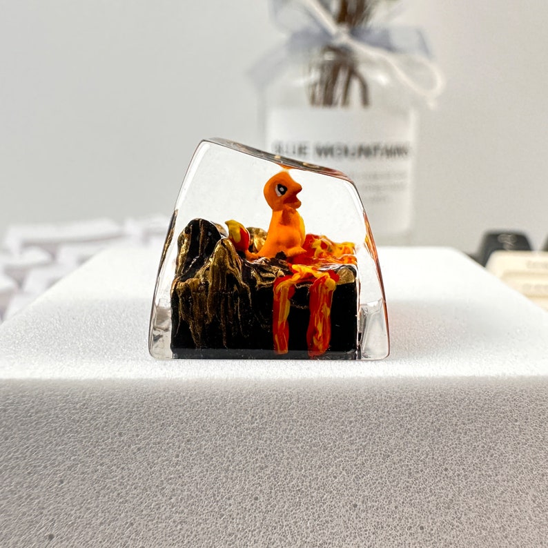 May include: A clear resin keycap featuring an orange cartoon character standing on a black and orange lava-like base.