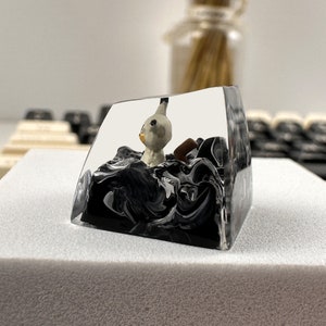 K&ouml;nnte beinhalten: Ein transparenter Keycap aus Harz mit einem schwarzen und wei&szlig;en Wirbelmuster. Im Inneren des Keycaps befindet sich eine kleine wei&szlig;e Figur eines Wesens mit langem Hals und rundem Kopf.