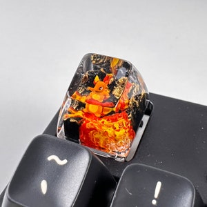 May include: A clear resin keycap with a black and gold background and a cartoon image of a fire-type Pokemon. The keycap is on a black keyboard.