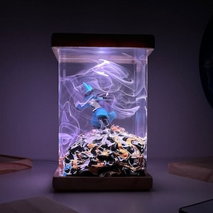 May include: A blue and white figurine of a character from a video game is encased in a clear resin cube with a swirling white design. The cube is lit from below with a purple light and sits on a wooden base.