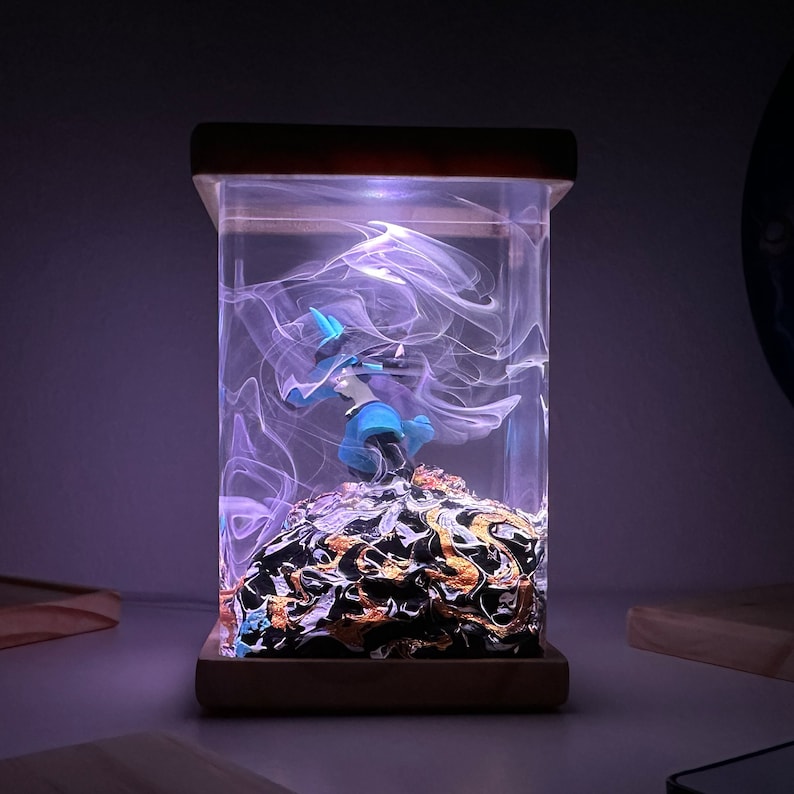 May include: A blue and white figurine of a character from a video game is encased in a clear resin cube with a swirling white design. The cube is lit from below with a purple light and sits on a wooden base.
