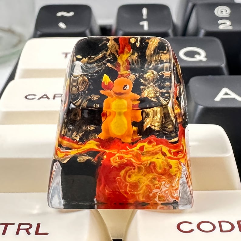 May include: A clear resin keycap with a black and gold background and a red and yellow flame design. A small orange and yellow cartoon character is in the center of the keycap.