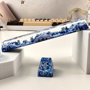 May include: A set of four blue and white resin keyboard wrist rests. The rests are shaped like waves and feature small figurines of penguins. The rests are displayed on a white surface.