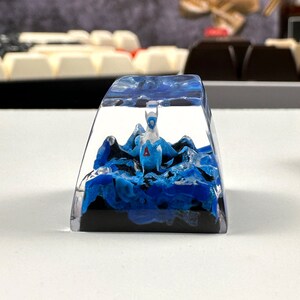 Pode incluir: Um keycap de resina transparente com um design de redemoinho azul e preto. Uma figurinha branca de uma criatura m&iacute;tica est&aacute; dentro do keycap.