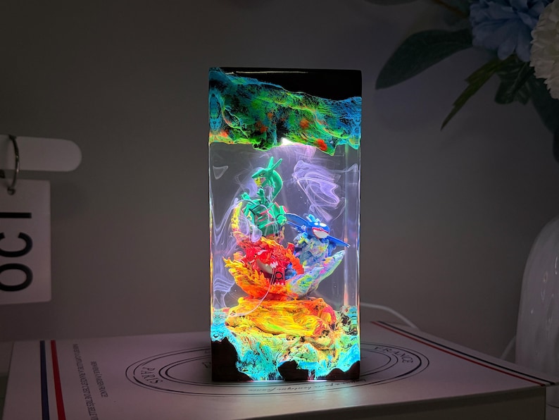 K&ouml;nnte beinhalten: Eine rechteckige Harzlampe mit einem klaren Mittelteil und interner LED-Beleuchtung. Im Inneren befinden sich drei Miniaturfiguren in Gr&uuml;n, Rot und Blau auf einem strukturierten Sockel. Die Kanten haben eine dunkle, marmorierte Oberfl&auml;che.