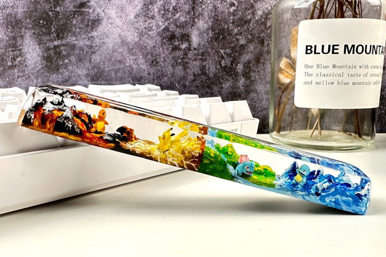 May include: A clear resin keyboard wrist rest with four different colored sections, each featuring a different Pokemon character. The sections are orange, yellow, green, and blue.