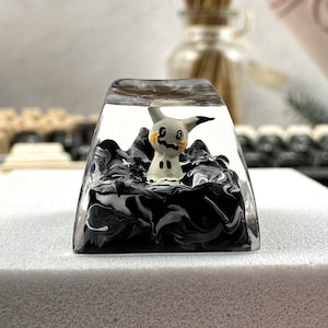 K&ouml;nnte beinhalten: Ein transparenter Keycap aus Harz mit einem schwarzen und wei&szlig;en Marmormuster. Im Inneren des Keycaps befindet sich eine kleine wei&szlig;e Figur eines Pok&eacute;mon-Charakters.