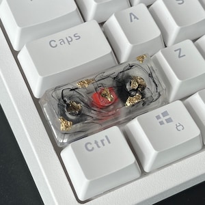 K&ouml;nnte beinhalten: Eine transparente Harztastenkappe mit schwarzen Wirbeln und goldenen Flocken. Die Tastenkappe hat einen roten Schalter im Inneren. Die Tastenkappe befindet sich auf einer wei&szlig;en Tastatur.