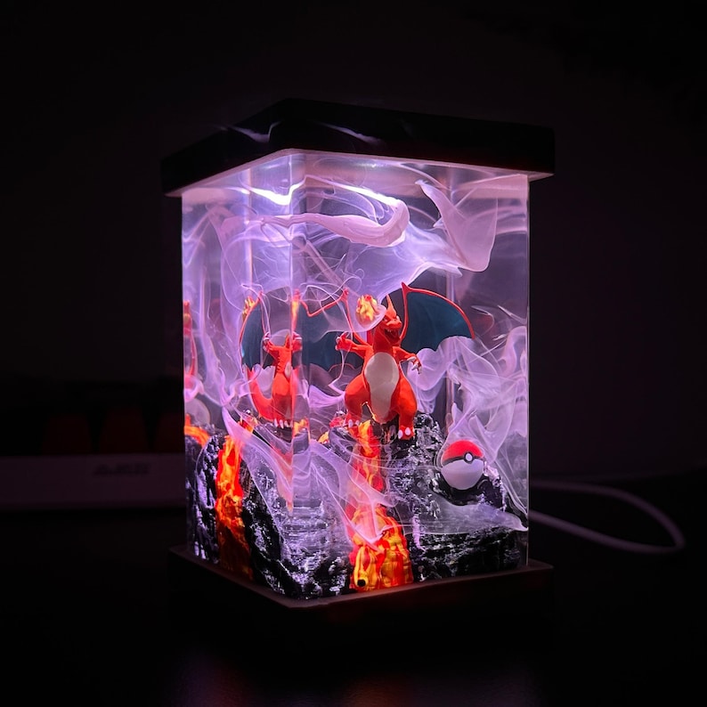 K&ouml;nnte beinhalten: Eine klare Glaslampe mit schwarzem Sockel zeigt eine 3D-Szene mit zwei roten und orangefarbenen Pok&eacute;mon-Charakteren, Charizard, in einer wirbelnden Rauchwolke mit einem roten und wei&szlig;en Pok&eacute;ball. Der Sockel der Lampe besteht aus schwarzem Harz und hat eine strukturierte Oberfl&auml;che.