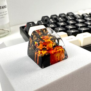 May include: A clear resin keycap with a black and orange design. The keycap features a small orange figurine inside, resembling a cartoon character. The keycap is sitting on a white surface.