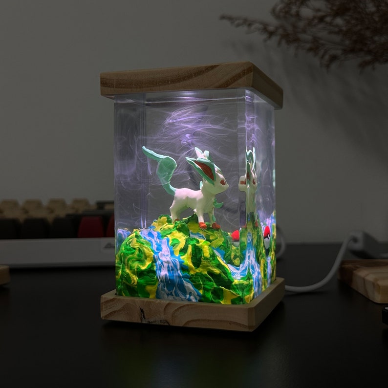 May include: A wooden lamp base with a clear resin cube containing a green and white figurine of a Pokemon character, a green and blue landscape, and a red and white Pokeball. The resin cube is illuminated from the inside with a white light.