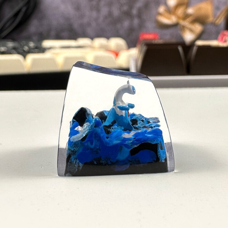 Pode incluir: Um keycap de resina transparente com um design de redemoinho azul e preto. Uma figura branca de uma criatura m&iacute;tica est&aacute; de p&eacute; na resina.