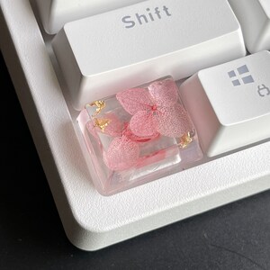 K&ouml;nnte beinhalten: Ein einzigartiger Tastenkappen f&uuml;r eine Tastatur mit einer rosa Blume, die in klarem Harz mit goldenen Flocken eingebettet ist.