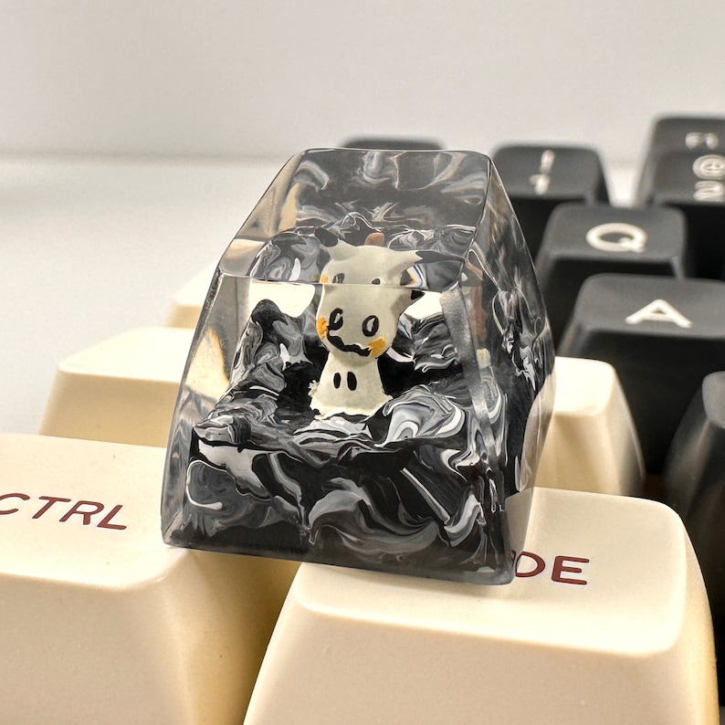 K&ouml;nnte beinhalten: Eine transparente Harztastenkappe mit einem schwarzen und wei&szlig;en Wirbelmuster. Eine kleine wei&szlig;e Figur eines Comic-Charakters befindet sich in der Tastenkappe.