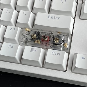 K&ouml;nnte beinhalten: Eine transparente Harz-Tastenkappe mit schwarzen Wirbeln, goldenen Flocken und roten und silbernen Akzenten. Die Tastenkappe ist f&uuml;r eine Standard-mechanische Tastatur ausgelegt.