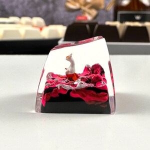 Pode incluir: Um keycap de acr&iacute;lico transparente com um design abstrato em rosa e preto e uma pequena figura branca e laranja dentro.