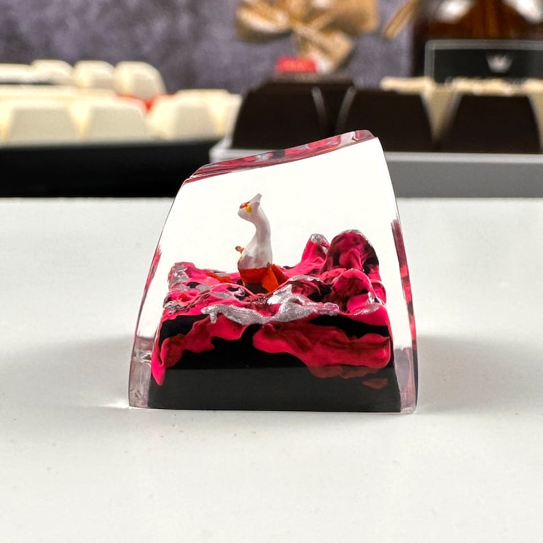 Pode incluir: Um keycap de acr&iacute;lico transparente com um design abstrato em rosa e preto e uma pequena figura branca e laranja dentro.