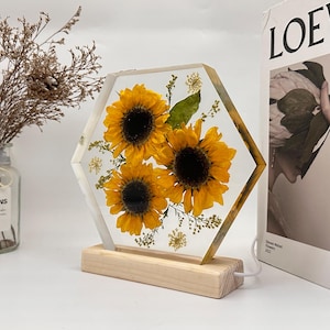 Pu&ograve; includere: Un blocco di resina trasparente esagonale contenente tre girasoli gialli pressati e piccolo fogliame verde, montato su una base rettangolare in legno chiaro. L'oggetto presenta un cavo di alimentazione bianco integrato sul lato del supporto.