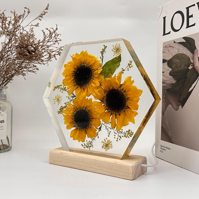 Pu&ograve; includere: Un blocco di resina trasparente esagonale contenente tre girasoli gialli pressati e piccolo fogliame verde, montato su una base rettangolare in legno chiaro. L'oggetto presenta un cavo di alimentazione bianco integrato sul lato del supporto.
