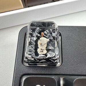 K&ouml;nnte beinhalten: Eine schwarz-wei&szlig;e marmorierte Tastenkappe mit einer kleinen wei&szlig;en Schneemannfigur im Inneren. Die Tastenkappe ist aus klarem Harz gefertigt und hat eine gl&auml;nzende Oberfl&auml;che.