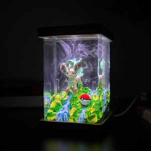 May include: A clear glass cube with a green and blue resin base, featuring a white and green Pokemon figurine and a red and white Pokeball. The cube is illuminated from within, creating a glowing effect.