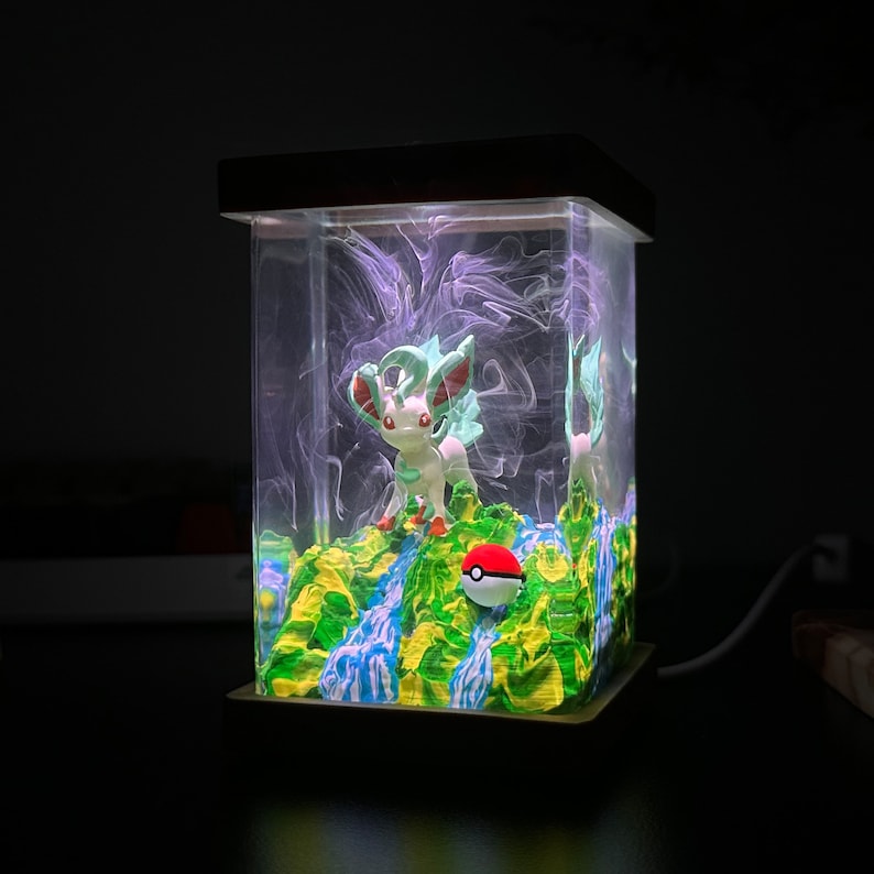 May include: A clear glass cube with a green and blue resin base, featuring a white and green Pokemon figurine and a red and white Pokeball. The cube is illuminated from within, creating a glowing effect.