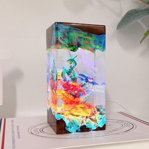K&ouml;nnte beinhalten: Eine rechteckige Lampe aus Harz und Holz mit einem klaren, beleuchteten Zentrum, das Miniaturfiguren von gr&uuml;nen, roten und blauen Kreaturen enth&auml;lt. Der Sockel und die Oberseite bestehen aus dunklem Holz mit leuchtenden Farben im Harz.