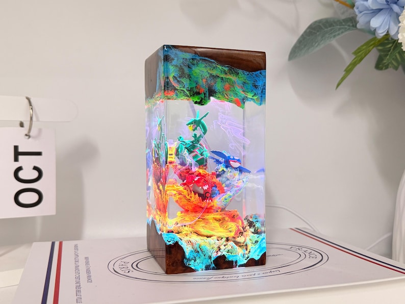 K&ouml;nnte beinhalten: Eine rechteckige Lampe aus Harz und Holz mit einem klaren, beleuchteten Zentrum, das Miniaturfiguren von gr&uuml;nen, roten und blauen Kreaturen enth&auml;lt. Der Sockel und die Oberseite bestehen aus dunklem Holz mit leuchtenden Farben im Harz.