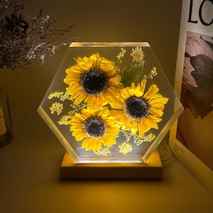 Pu&ograve; includere: Un blocco esagonale in resina trasparente contenente tre girasoli gialli conservati e piccoli fiori verdi. Il pezzo in resina &egrave; montato su una base rettangolare in legno chiaro che illumina i fiori dal basso con una luce calda.