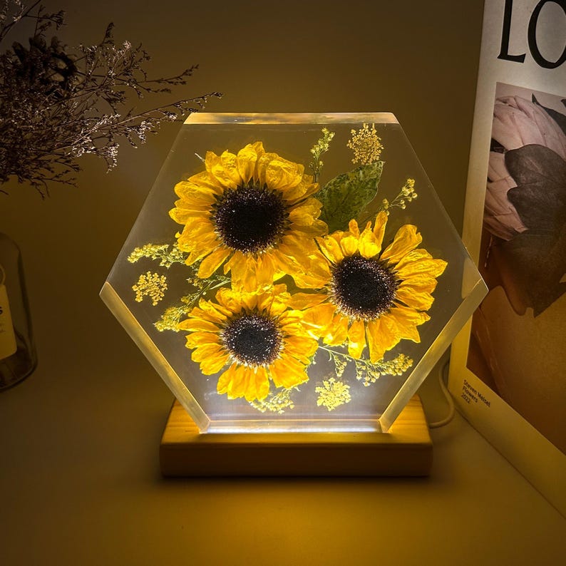 Pu&ograve; includere: Un blocco esagonale in resina trasparente contenente tre girasoli gialli conservati e piccoli fiori verdi. Il pezzo in resina &egrave; montato su una base rettangolare in legno chiaro che illumina i fiori dal basso con una luce calda.