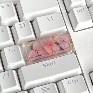 K&ouml;nnte beinhalten: Eine wei&szlig;e Tastatur mit einer rosafarbenen und goldenen Harztastatur mit gepressten Blumen. Die Tastatur ist mit "Shift" beschriftet.
